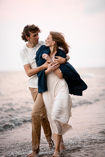 Husband and his wife in love are walking on the water along the seashore, the husband hugs his wife tightly from the side, looking into her eyes