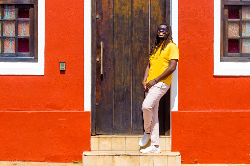handsome black man with afro hair wears sunglasses, yellow shirt and white pants under red facade of old house. smiles at the camera while looking up