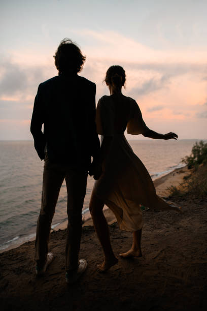 Young and slender newlyweds stand on the sandy seashore holding hands against the background of the sea and sunset sky