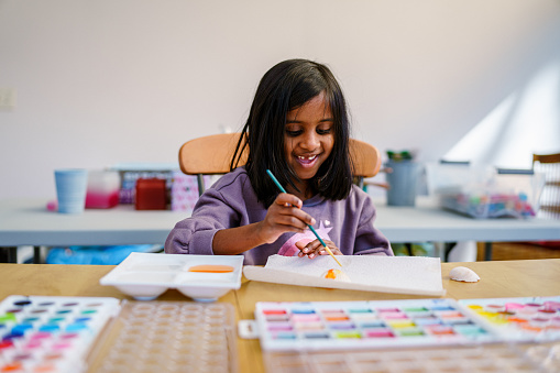 A cute elementary school age girl of Indian descent smiles while sitting at a table, painting while enjoying arts and crafts on a day off at home.