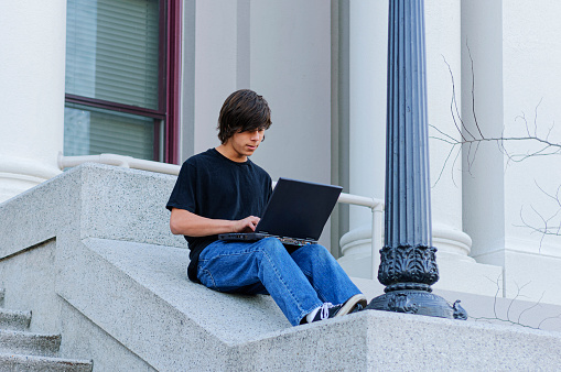 One teenage Latino boy sitting on the steps outside of school, working on his computer.