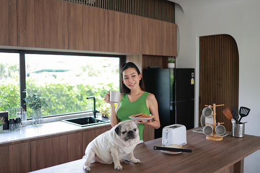 A woman is standing in a kitchen with a pug dog. She is holding a plate of food and a cup of coffee. The kitchen is well-equipped with a toaster, a sink, and a refrigerator