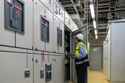 Professional Asian man engineer in safety uniform working on digital tablet at factory server electric control panel room. Industrial technician worker maintenance power system at manufacturing plant room.