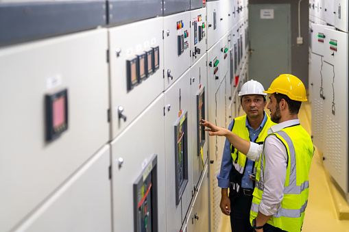 Two Professional electrical engineer in safety uniform working at factory server electric control panel room. Industrial technician worker maintenance checking power system at manufacturing plant room.