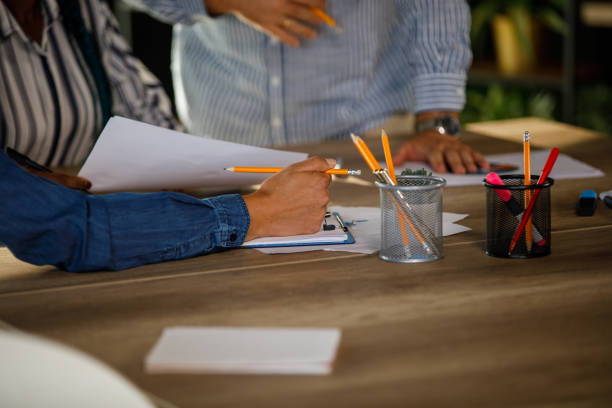 Candid shot of two diverse businesswomen and their male colleague, standing by the conference table, reviewing documents and chatting about strategies during a project briefing.