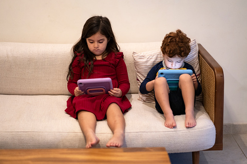 Two siblings 3 and 5 years old sitting on a couch on the living room holding each a digital tablet in their hands watching the illuminated screens.