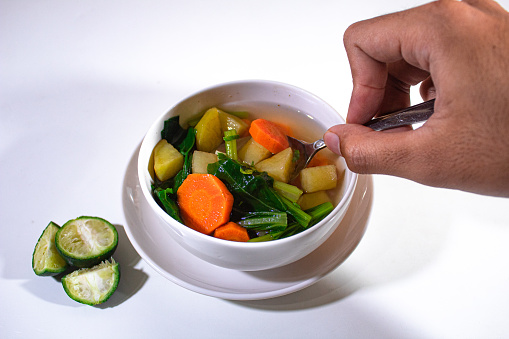 a porcelain bowl containing carrot, potato and mustard greens soup, served on a plastic plate decorated with lime and the right hand spooning the soup