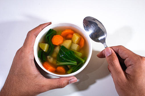 a porcelain bowl held in the left hand, containing vegetable soup and the right hand holding a kitchen spoon