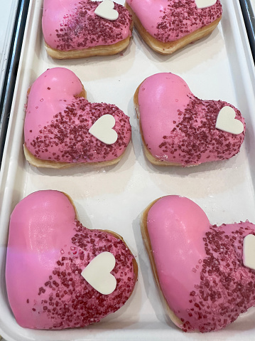 Stock photo showing close-up, elevated view of rows of pink fondant iced heart doughnuts on white bakery tray.  The iced fried dough desserts are decorated with red sugar sprinkles and white chocolate hearts.