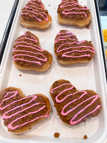 Stock photo showing close-up, elevated view of rows of glazed heart doughnuts on white bakery tray.  The iced fried dough desserts are decorated with biscuit crumbs and pink icing pattern.