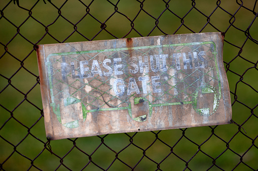 View of an old faded sign on a sheet of tin attached to a rusty chain wire fence asking to please shut the gate
