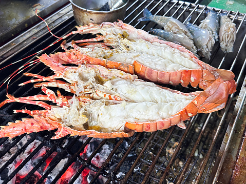 Stock photo showing close-up, elevated view of ash and flaming coals burning on a barbecue, topped with metal grate, at an outdoor beach restaurant, which has been lit in preparation of cooking seafood.
