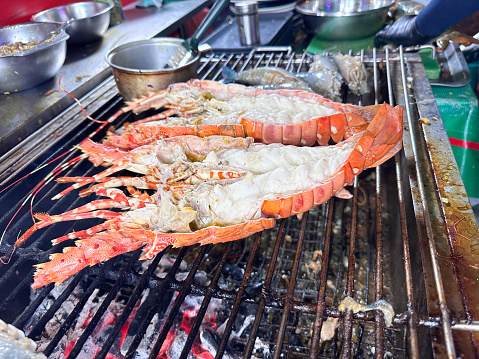 Stock photo showing close-up view of ash and flaming coals burning on a barbecue, topped with metal grate, at an outdoor beach restaurant, which has been lit in preparation of cooking seafood.
