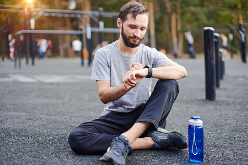 Young man sitting on the ground in the park getting ready for his outdoor workout, setting up smart watch mobile tracking application
