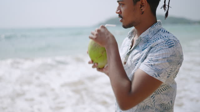 Man sips fresh coconut on tropical beach, sea waves in background. Relaxed male enjoys natural drink, beach lifestyle. Tourist hydrates with fruit on sunny shore. Healthy, vacation, refreshment theme.