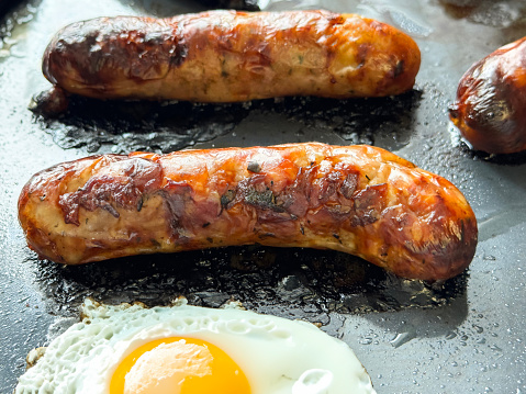 Stock photo showing close-up  view of a metal serving tray containing sausages and a sunny-side-up fried egg and grilled tomatoes for a breakfast self-service buffet at a hotel, to be eaten as part of a greasy and filling meal.