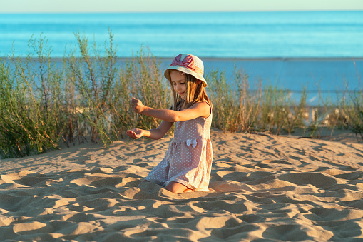 Happy little girl plays with sand on beach on background at blue sea at sunset. Summer vacation, travel, holiday. Child outdoors