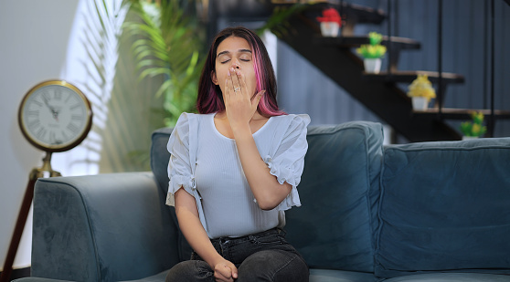 Indian sleepy bored young girl sitting on sofa yawning covering mouth with hand feeling tired drowsy at home. Sleepless alone lazy female sighing falling asleep suffer lack of sleep indoor house