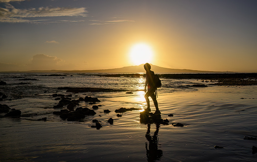 Woman walking on rocks at Milford Beach. Sunrise over the Rangitoto Island. Auckland.