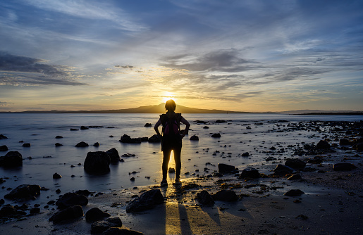 Woman standing among rocks at Milford Beach. Sunrise over the Rangitoto Island. Auckland.