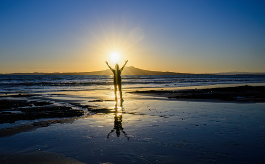 Woman raises arms towards the rising sun. Sunrise over the Rangitoto Island. Milford Beach. Auckland.