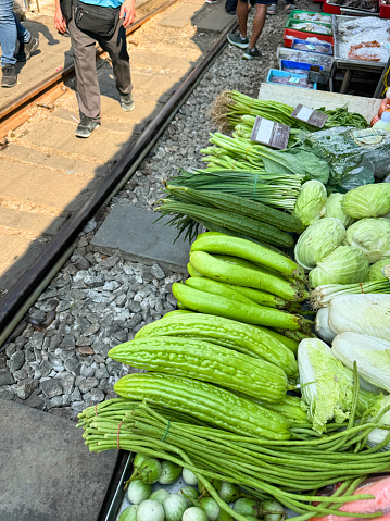 Stock photo showing a close-up, elevated view of fruit and vegetables being sold by market traders from outdoor ground stall display at the railway track at Maeklong Market, Bangkok, Thailand. Produce includes spring onions, cauliflowers, tomatoes, onions, cauliflowers, cabbages, gourds and herbs.