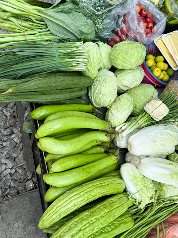 Stock photo showing a close-up, elevated view of fruit and vegetables being sold by market traders from outdoor ground stall display at the railway track at Maeklong Market, Bangkok, Thailand. Produce includes spring onions, cauliflowers, tomatoes, onions, cauliflowers, cabbages, gourds and herbs.