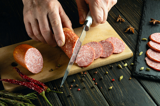 Slicing meat veal sausage with a knife in the hand of a cook on the kitchen table. Low key concept for table setting in a restaurant.