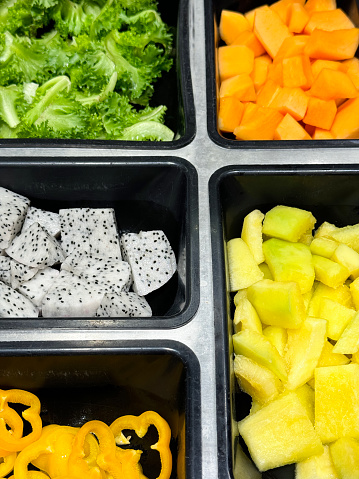 Stock photo showing close-up, elevated view of yellow bell pepper slices, dragon fruit (pitaya) pieces, lettuce leaves, cubed pineapple and mango in plastic serving trays as part of hotel self service refrigerated buffet display.