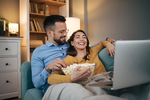A young couple in love is lying on the bed in their living room, enjoying watching a movie and eating popcorn.They laugh and have a great time
