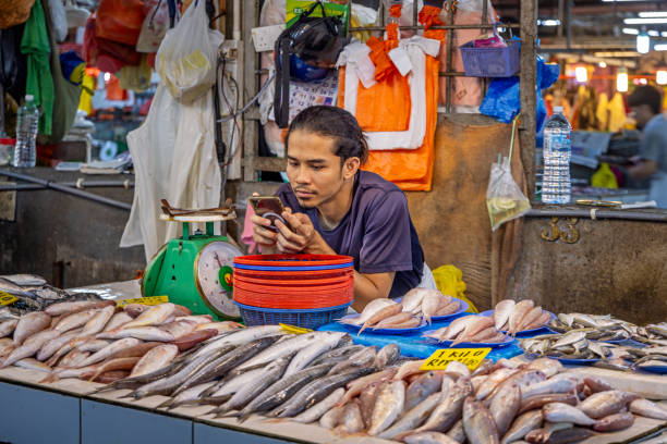 Chow Kit market, Kuala Lumpur, Malaysia - January 10th 2024:  Male fishmonger having a quiet moment with his mobile phone  at a stall in the famous food market in the center of the capital