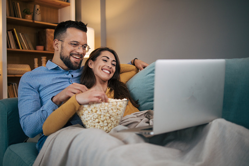 A young couple in love is lying on the bed in their living room, enjoying watching a movie and eating popcorn.They laugh and have a great time