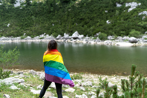 Unrecognizable woman on her back waves the gay rainbow flag with the mountain lake in the background