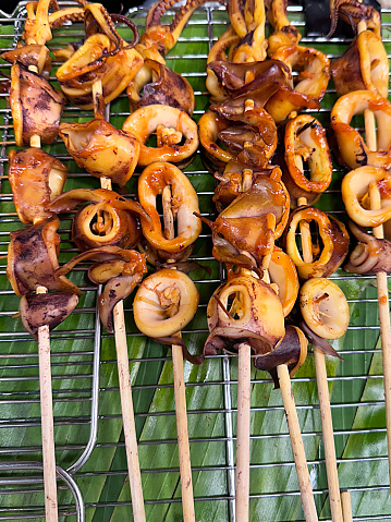 Stock photo showing close-up, elevated view of grilled, squid rings on bamboo skewers lying in rows on a metal grill rack over a layer of banana leaves.