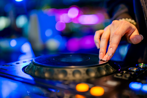 A skilled DJ’s hand deftly manipulates a turntable amidst vibrant club lights. The close-up view captures the DJ’s well-manicured nails as they work the turntable, part of a larger mixing console.