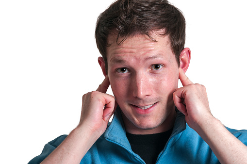 Frontal head portrait of a young brunette Caucasian man sticking his index fingers in his ears with an annoyed facial expression and looking at the camera isolated on white.