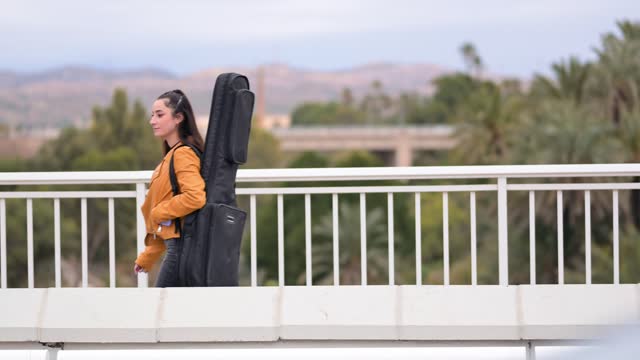 Young woman carrying guitar on her back walking across urban bridge