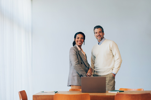 Two professional business colleagues sharing a pleasant moment, standing with a laptop in a bright, contemporary office space.