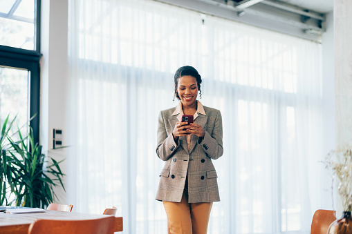 A young professional woman in a houndstooth blazer smiles at her smartphone, standing in a well-lit office space.