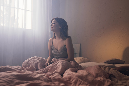 Young woman sitting on a bed, peaceful as morning light filters through the window. A moment of tranquility and introspection.