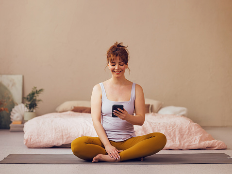Cheerful young woman sits on a yoga mat at home, browsing her smartphone with a relaxed, happy demeanor in a warmly lit bedroom.