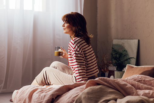 Serene young woman with coffee cup sitting on bed, basking in natural light from window in a cozy bedroom.