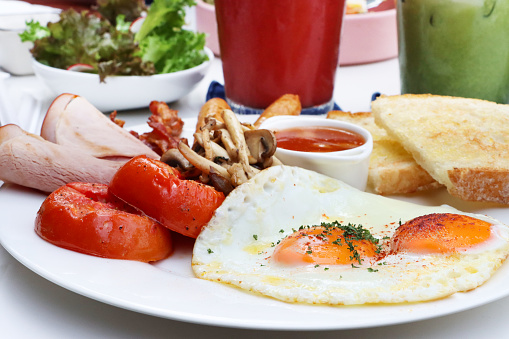 Stock photo showing close-up view of fried breakfast on white plate, slice of toasted white bread, two sunny-side-up fried eggs, ramekin of baked beans, ham slices, grilled tomatoes and mushrooms.