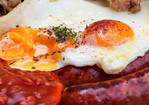 Stock photo showing close-up view of fried breakfast ingredients, baked beans, two sunny-side-up fried eggs, mushrooms and sausages.