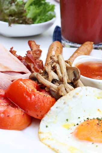 Stock photo showing close-up view of fried breakfast on white plate, slice of toasted white bread, two sunny-side-up fried eggs, ramekin of baked beans, ham slices, grilled tomatoes and mushrooms.