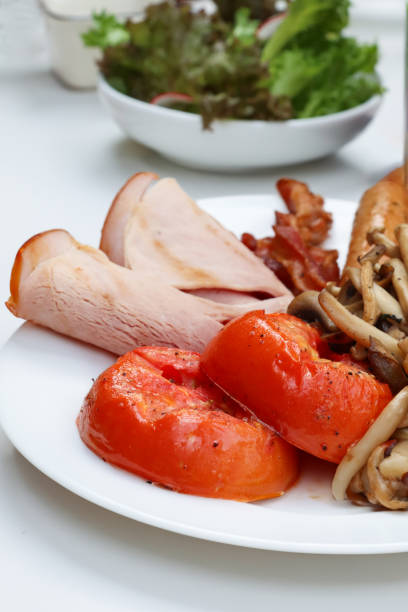 Stock photo showing close-up view of fried breakfast on white plate, ham slices, streaky bacon rashers, sausage, grilled tomato and mushrooms.