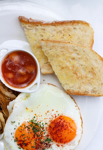 Stock photo showing close-up, elevated view of fried breakfast on white plate, slice of toasted white bread, two sunny-side-up fried eggs, ramekin of baked beans, pile of mushrooms.