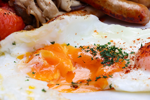 Stock photo showing close-up view of fried breakfast ingredients, grilled tomato, two sunny-side-up fried eggs, mushrooms and sausages.