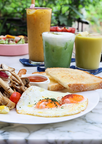 Stock photo showing close-up view of fried breakfast on white plate, slice of toasted white bread, sunny-side-up fried eggs, ramekin of baked beans, sausage, grilled tomato and mushrooms.