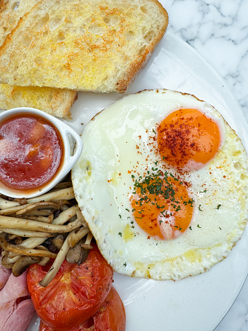 Stock photo showing close-up, elevated view of fried breakfast on white plate, slice of toasted white bread, two sunny-side-up fried eggs, ramekin of baked beans, ham slices, grilled tomatoes and mushrooms.